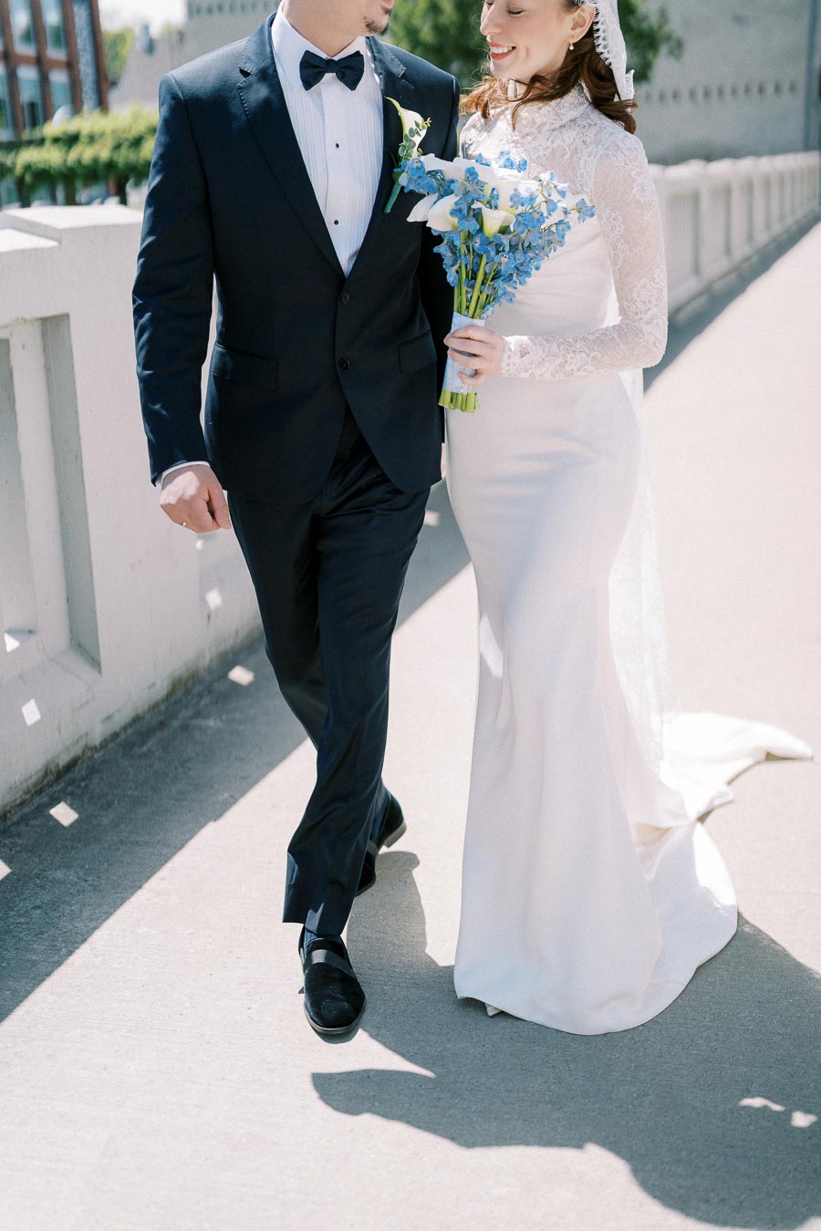 Bride and groom walking arm in arm on a sunny day, with the bride in an elegant lace wedding dress holding a bouquet of blue and white flowers, and the groom in a classic black tuxedo.
