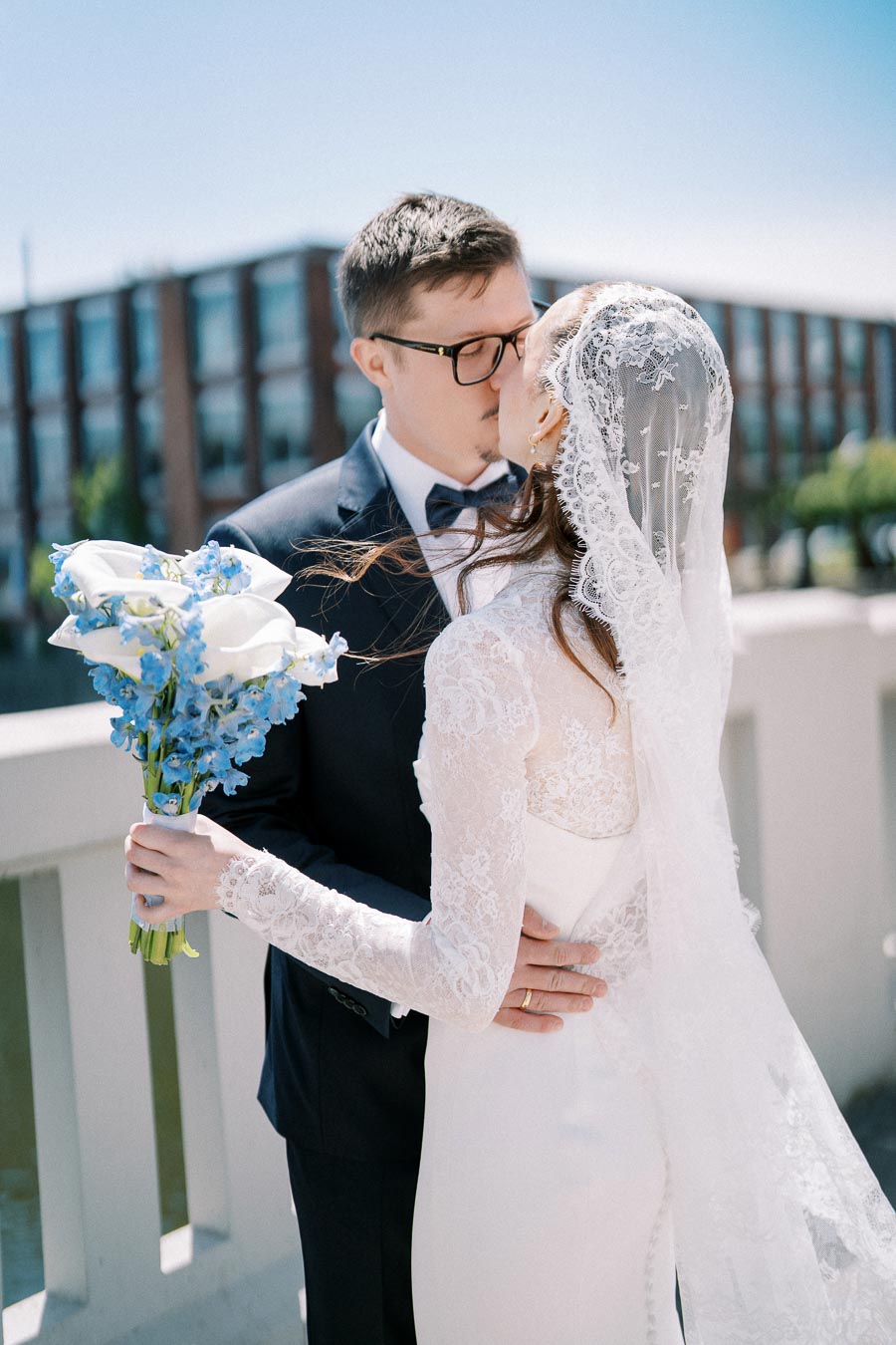 A bride and groom sharing a kiss outdoors on a sunny day, with the bride holding a bouquet of blue and white flowers, and both dressed in elegant wedding attire.