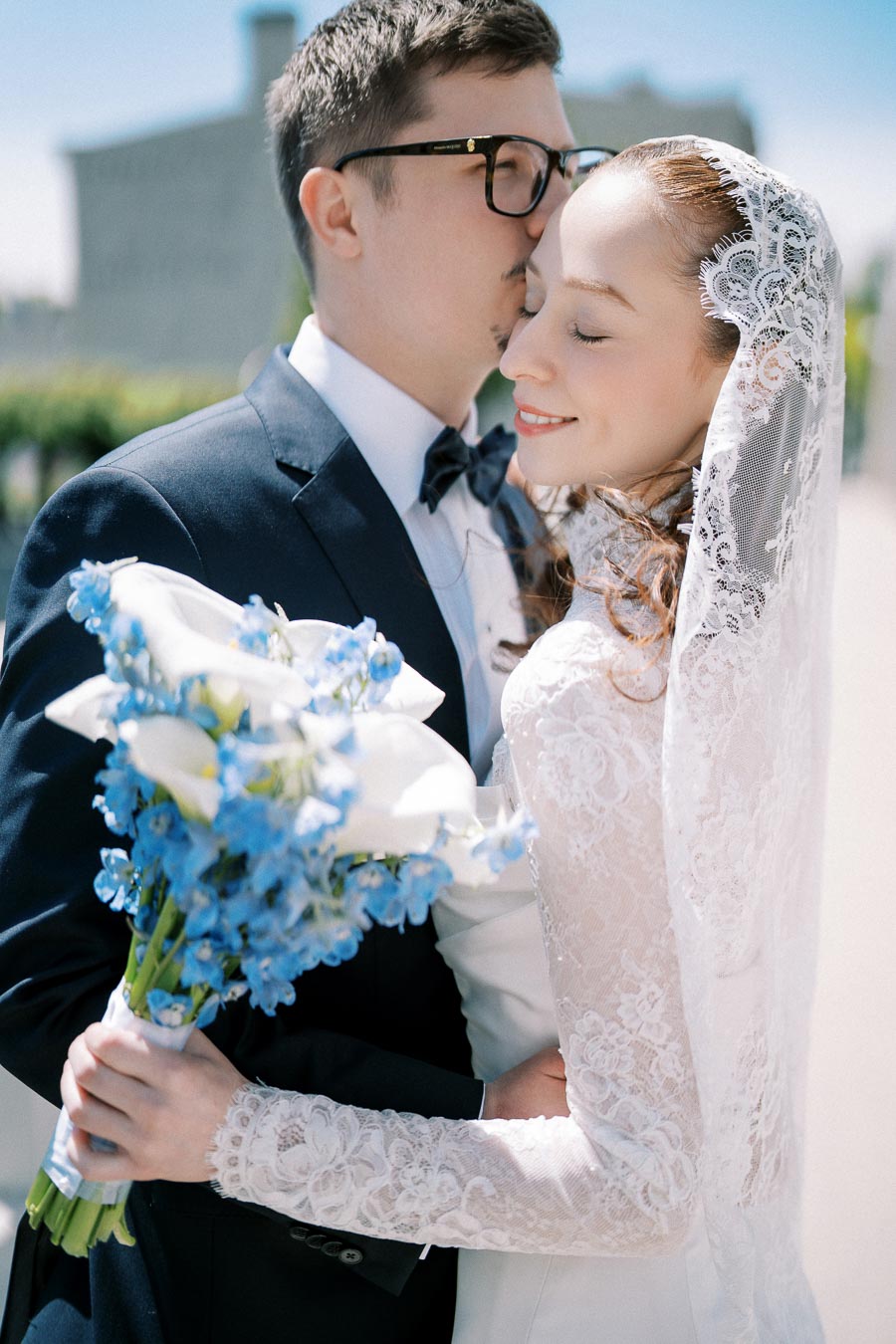 A groom in a navy blue suit kisses his bride on the forehead. The bride, in an elegant white lace dress and veil, holds a bouquet of blue and white flowers, smiling with closed eyes. The background is a sunny outdoor setting.