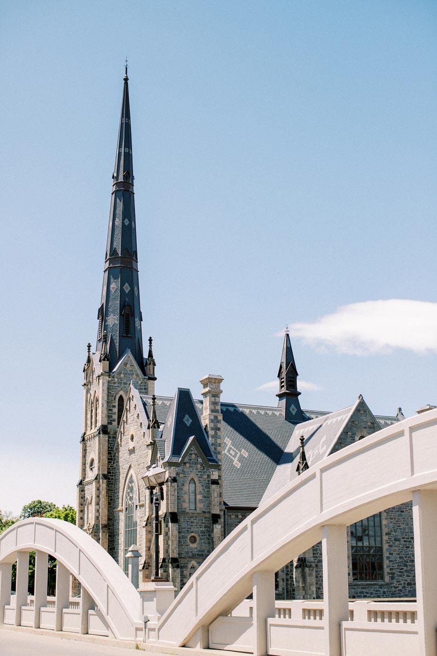Historic stone church with tall steeple and intricate architectural details behind an arched white bridge under a clear blue sky.