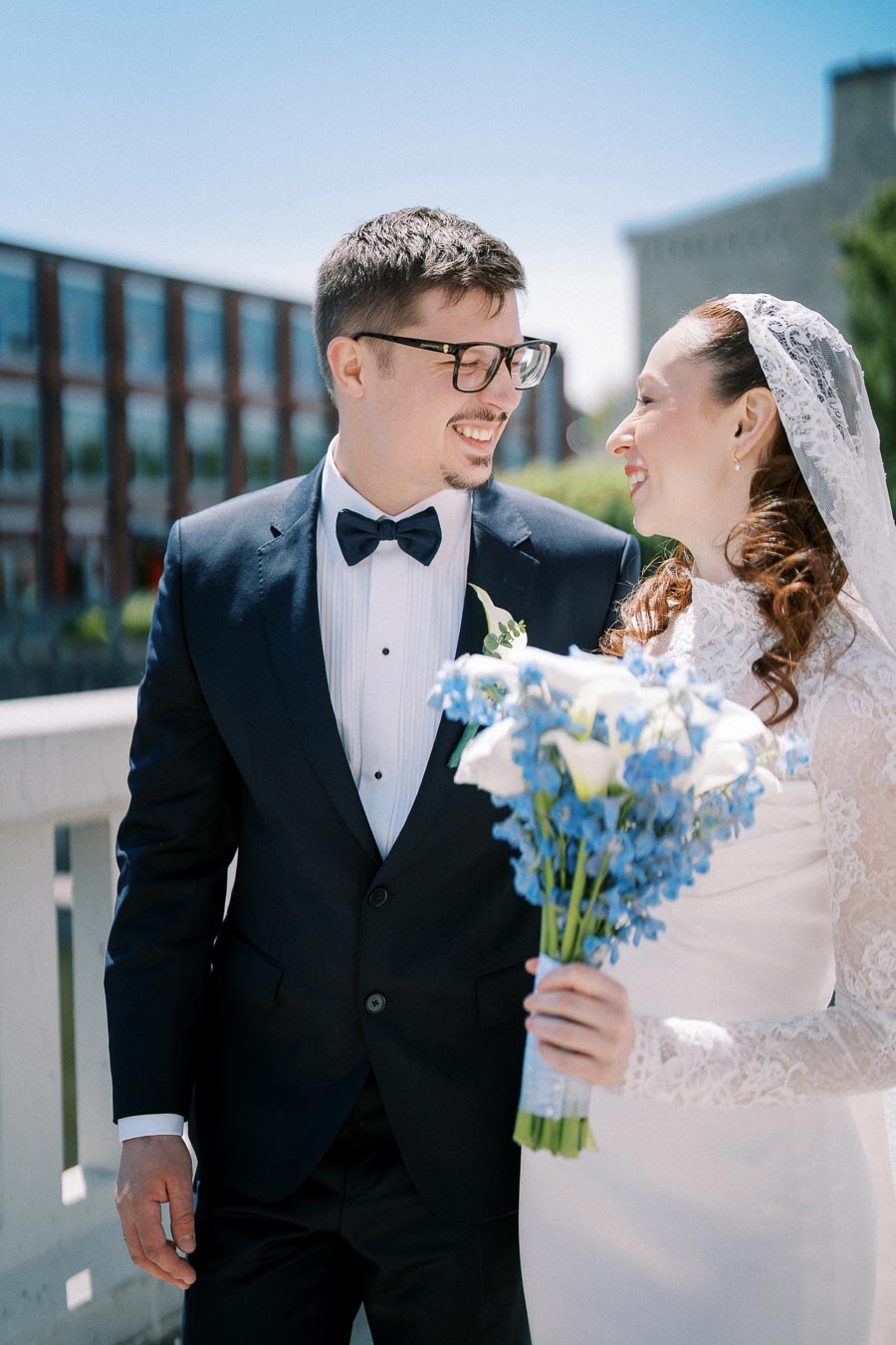 A bride and groom smiling at each other on a sunny day, with the bride holding a bouquet of blue flowers, both dressed in elegant wedding attire against an urban backdrop.