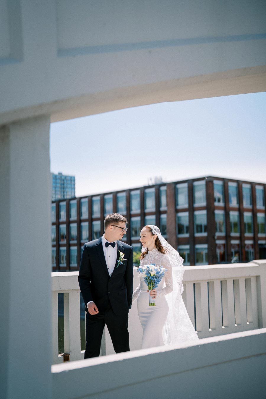 A bride and groom smiling at each other on a bridge, with a modern building in the background. The bride holds a bouquet of blue and white flowers, and they are both dressed in formal wedding attire under a clear blue sky.