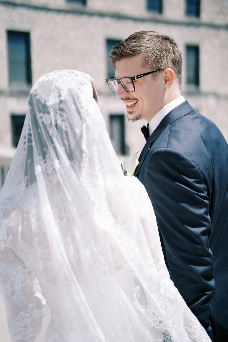 A bride and groom smiling at each other outdoors on their wedding day, with the groom in a navy suit and the bride wearing a white lace veil, set against a stone building background.