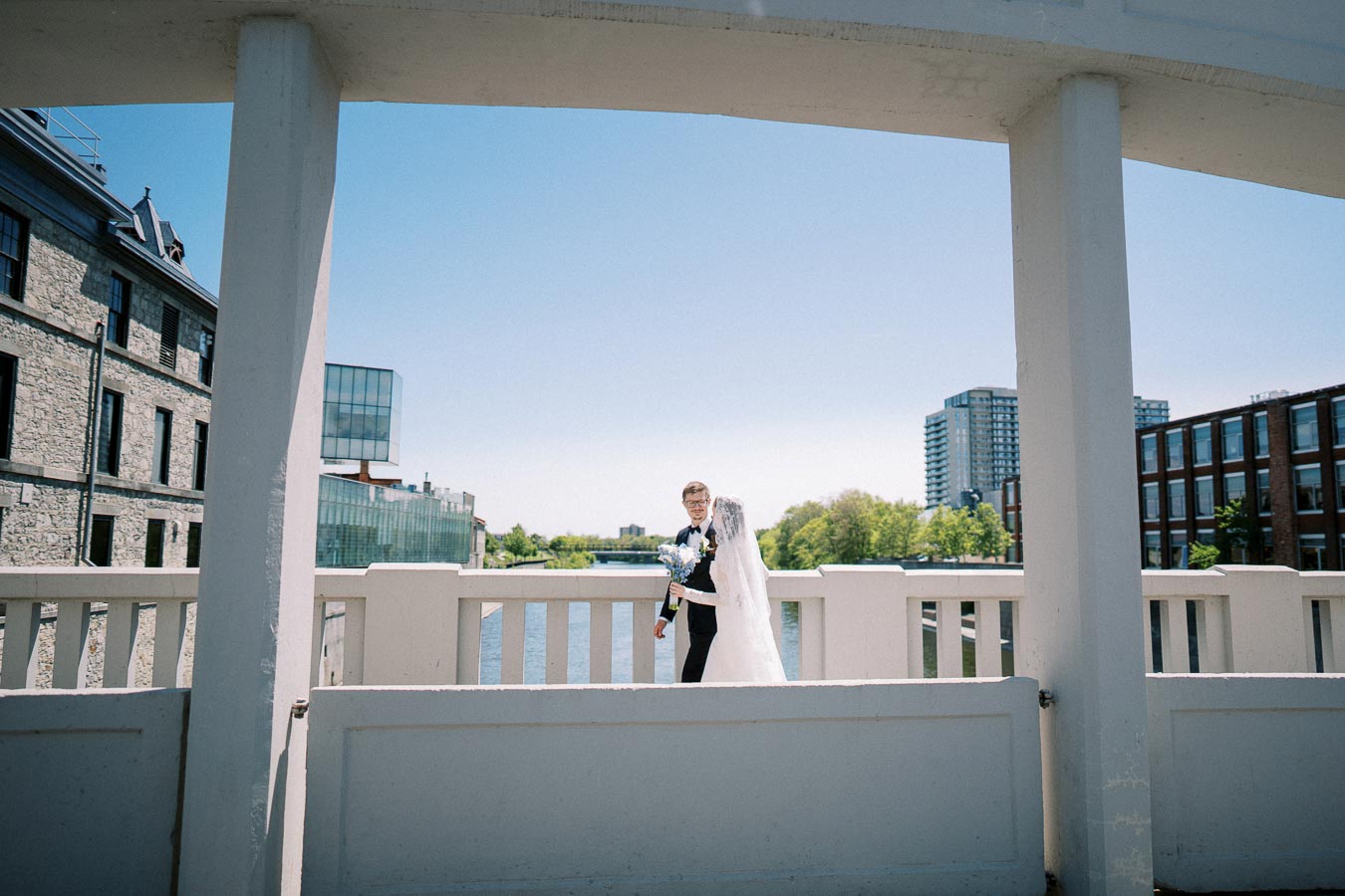 Wedding couple posing on a bridge with urban architecture and river in the background, featuring a groom in a suit and bride in a white gown, capturing a beautiful sunny day.