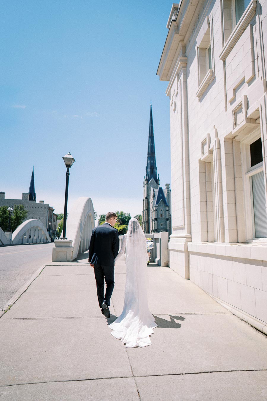 A bride and groom walking on a city sidewalk towards an ornate church on a clear day, with modern urban architecture and a bridge in the background.