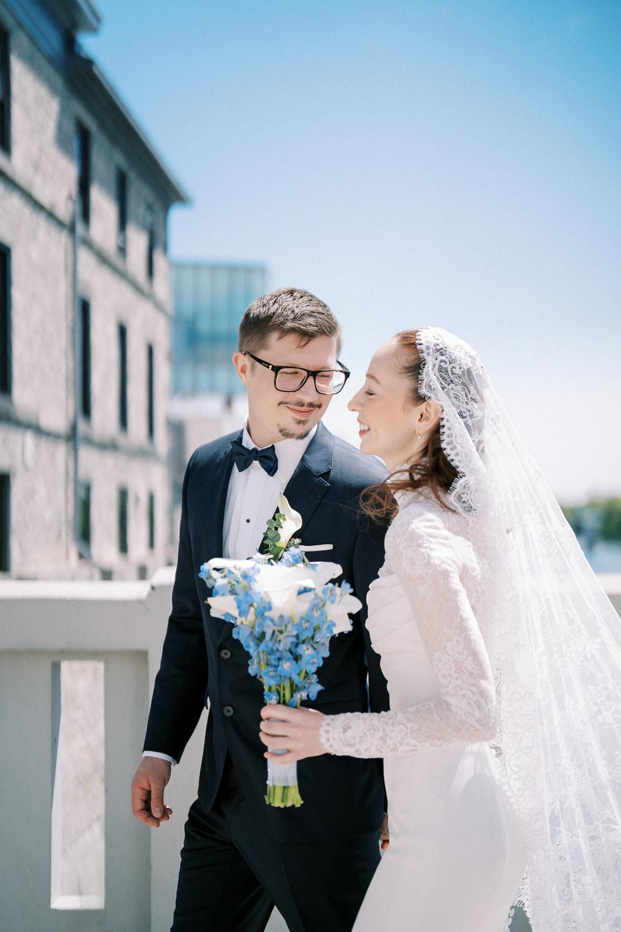 A bride and groom smiling at each other on their wedding day, with the bride holding a bouquet of blue and white flowers. The couple is standing outdoors near a building under a clear blue sky.