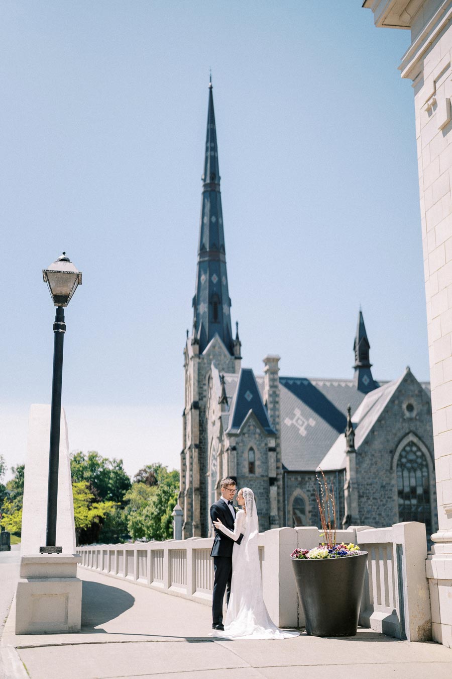 Wedding couple embracing on a sunny day in front of a historic church with a tall spire, surrounded by beautiful architecture and a vibrant planter with flowers.