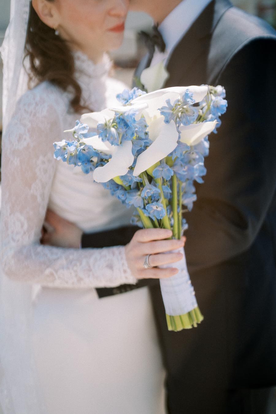 Elegant bride in lace wedding dress holding bouquet of white calla lilies and blue flowers, embracing groom in black suit and bow tie, capturing a romantic wedding moment.