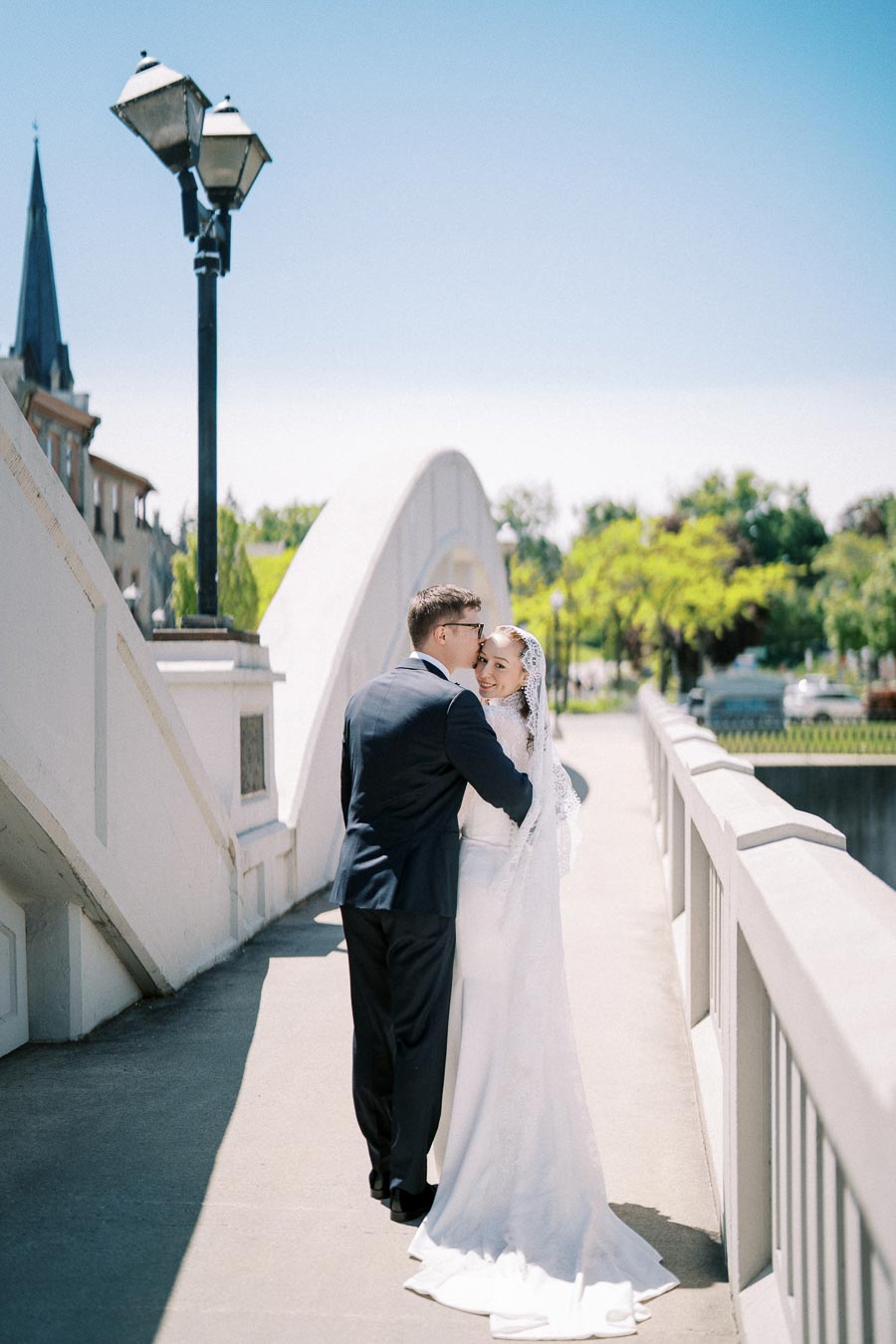 Elegant bride and groom embracing on a sunlit bridge, with a picturesque cityscape and clear blue sky in the background, capturing a romantic wedding moment.