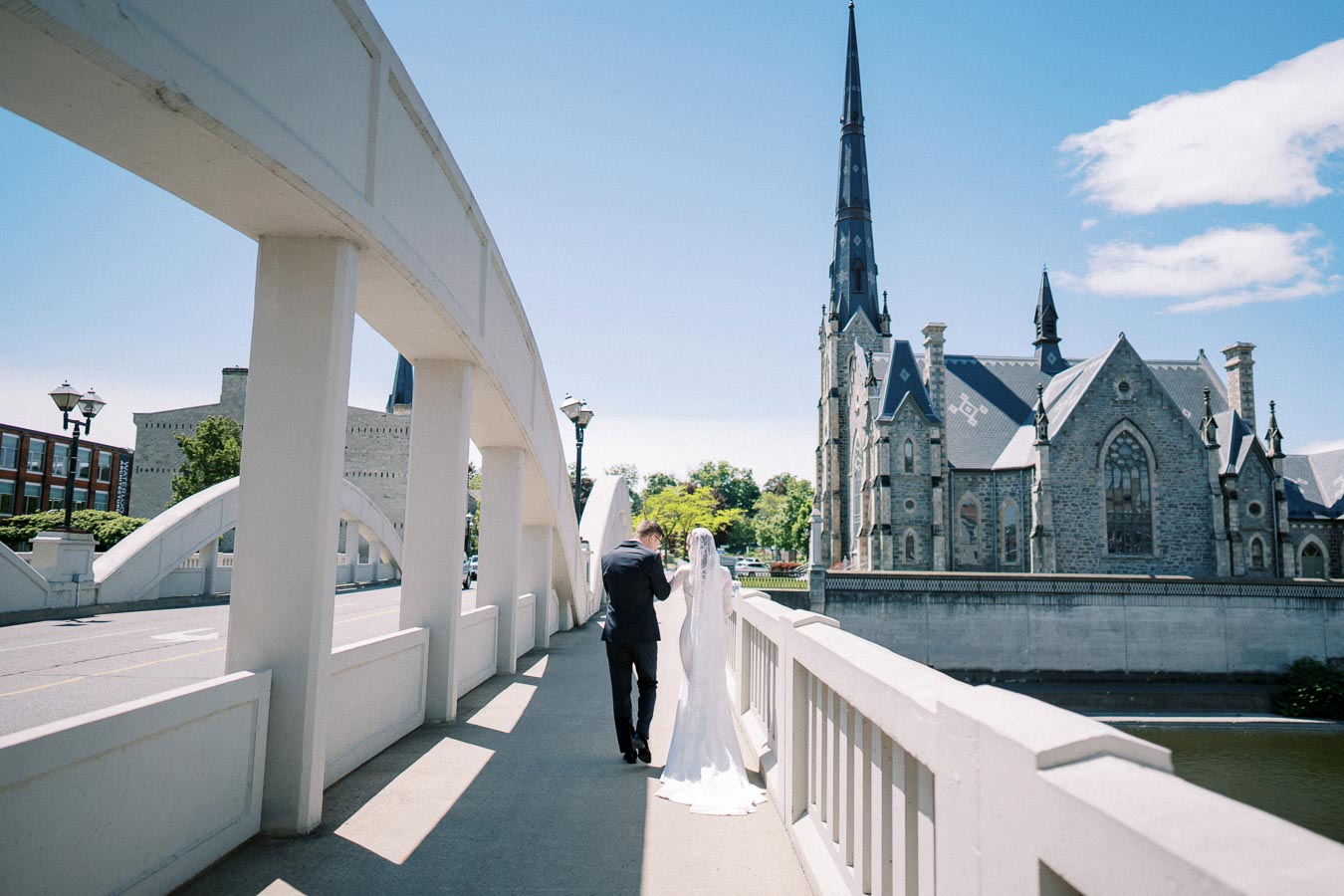 A bride and groom walk hand in hand across a white bridge with a historic stone church and clear blue sky in the background, creating a picturesque wedding scene.