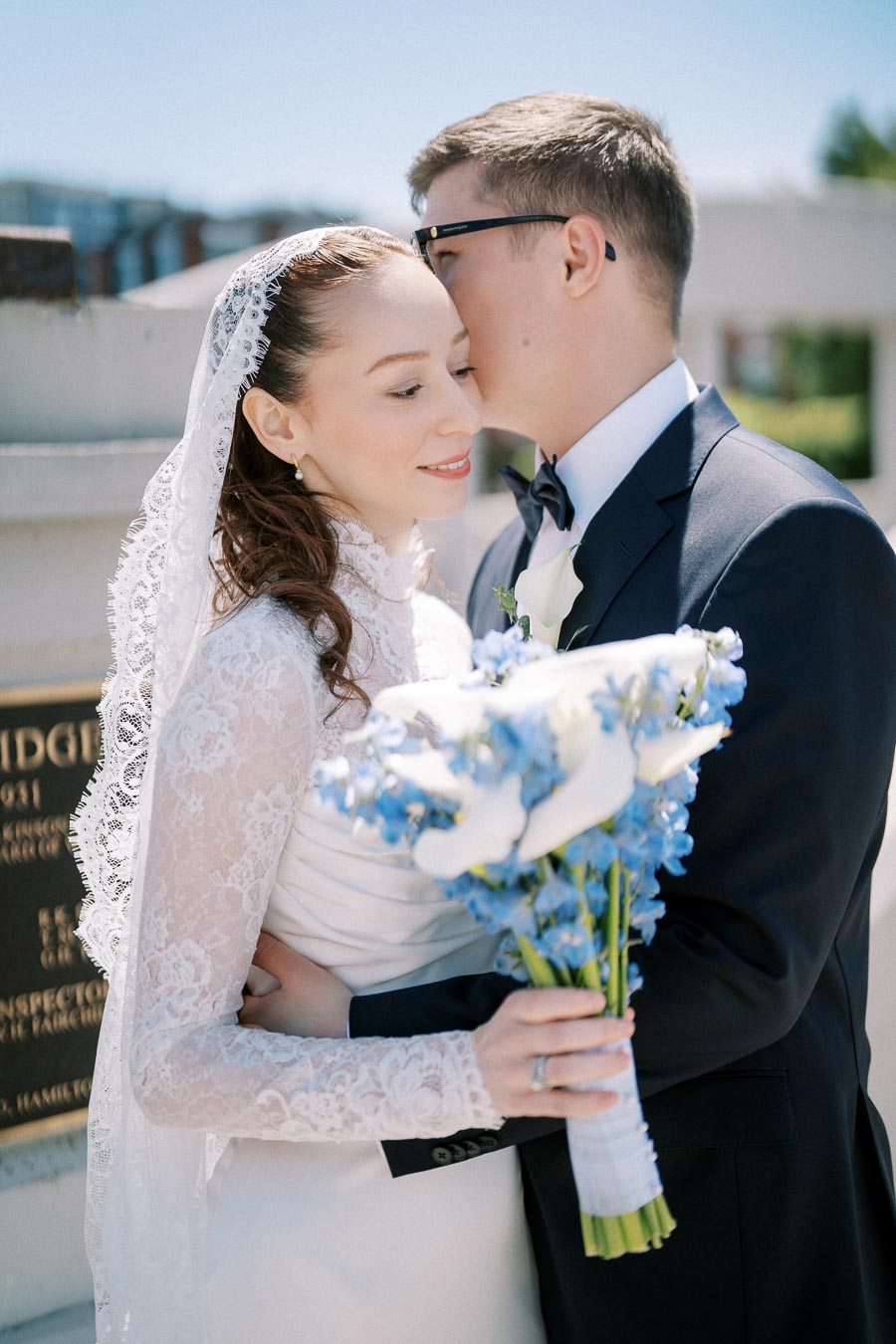 A bride and groom embrace in an outdoor setting; bride wearing a lace veil holds a bouquet of white and blue flowers, while the groom in a dark suit whispers in her ear, capturing a romantic wedding moment.
