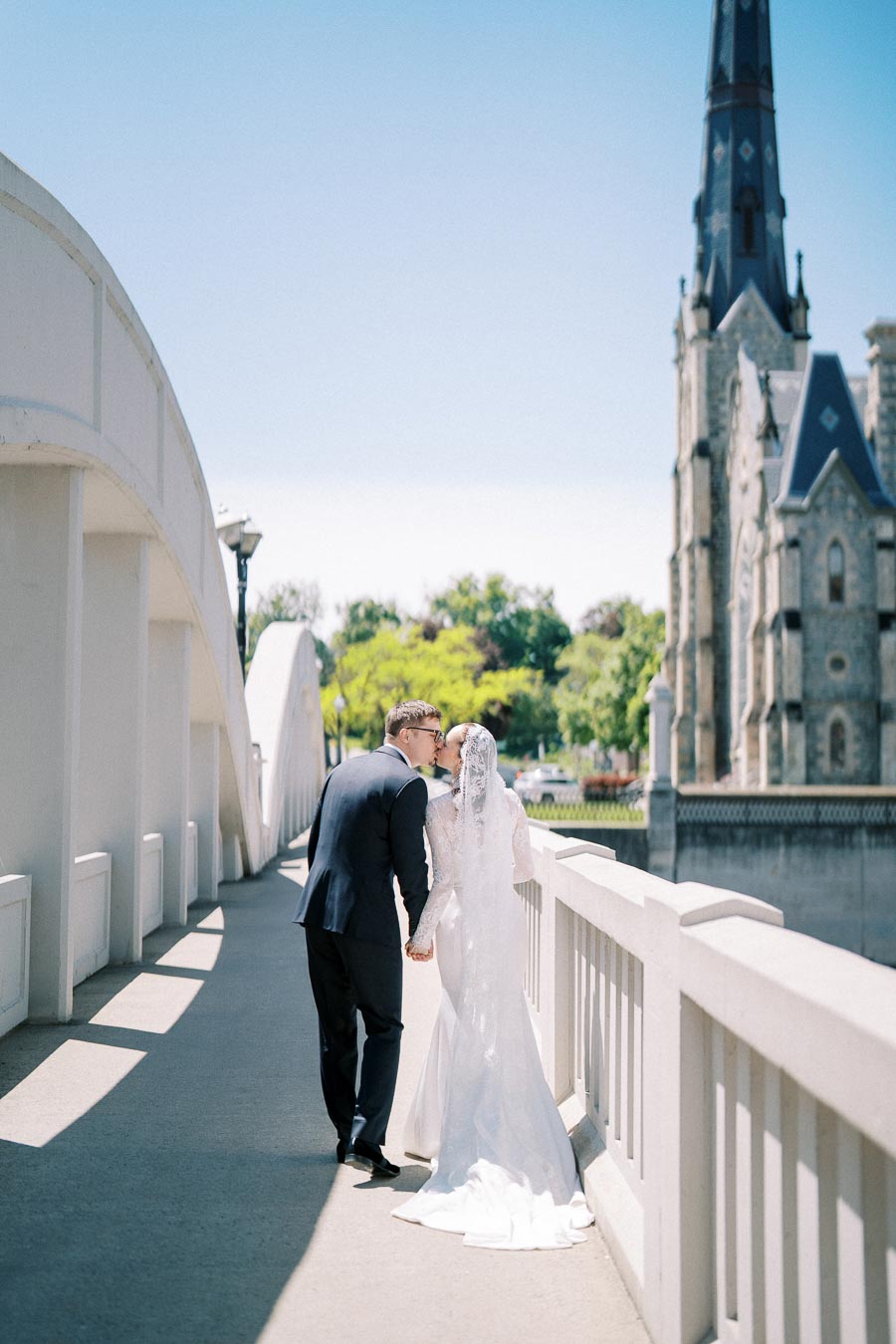 A bride and groom share a kiss on a bridge with a picturesque church in the background, under a clear blue sky.