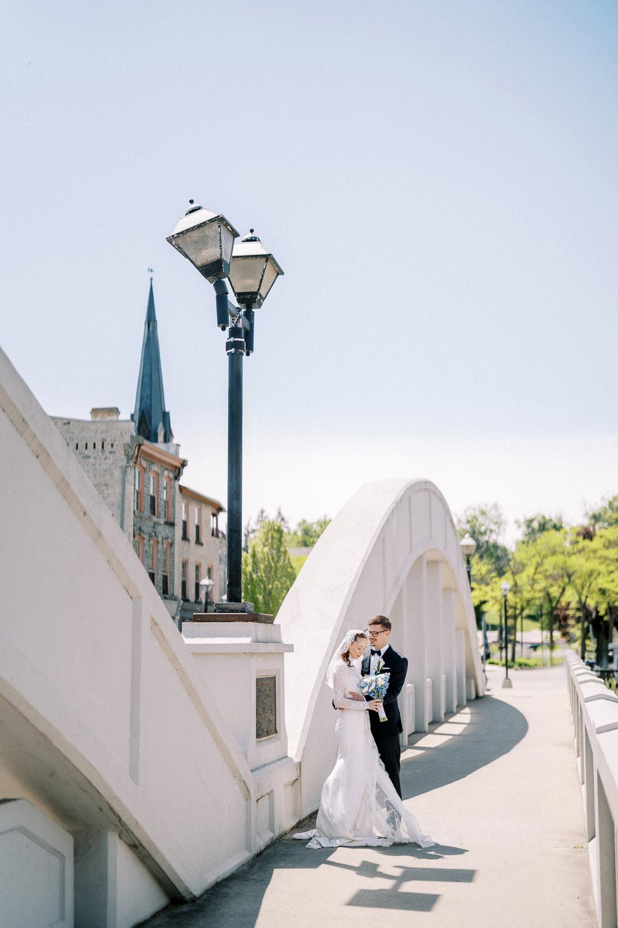 Wedding couple embracing on a white arched bridge, with a historic building and streetlamp in the background, under a clear blue sky.