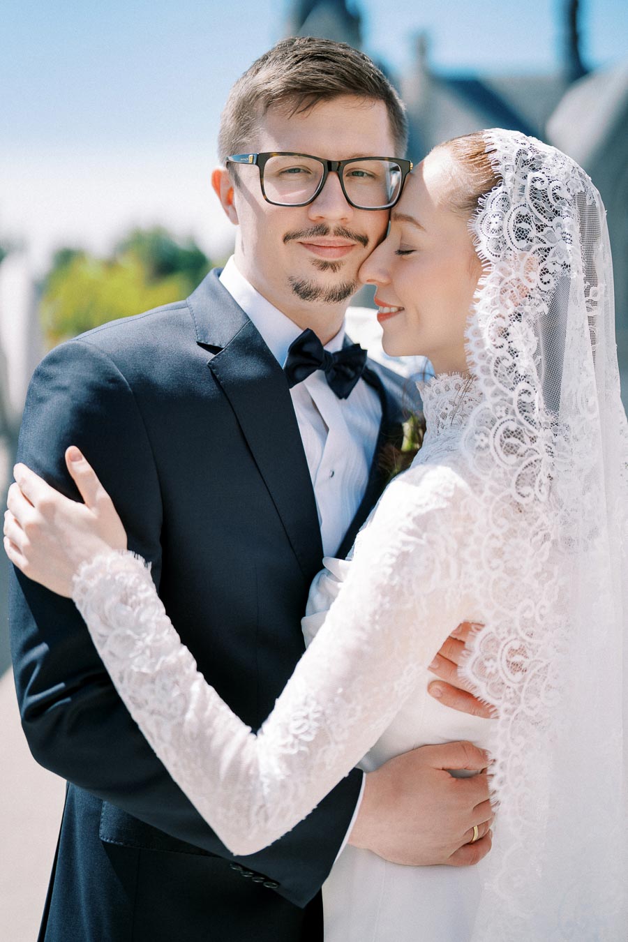 A bride in a lace veil embraces a groom in a black tuxedo and glasses, both smiling tenderly on their wedding day outdoors.