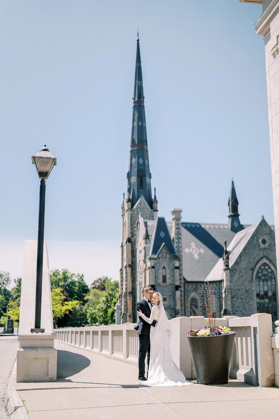 Wedding couple embracing on a sunny day in front of a historic church with a tall spire, surrounded by urban architecture and a decorative flower pot.