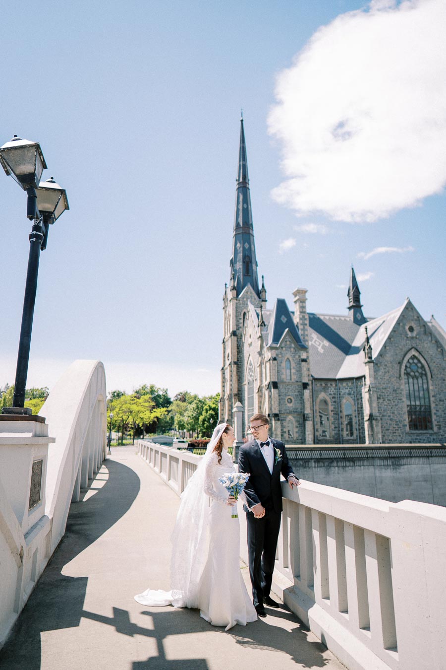 A bride and groom in wedding attire stand on a picturesque bridge in front of a historic stone church with a tall spire under a clear blue sky.