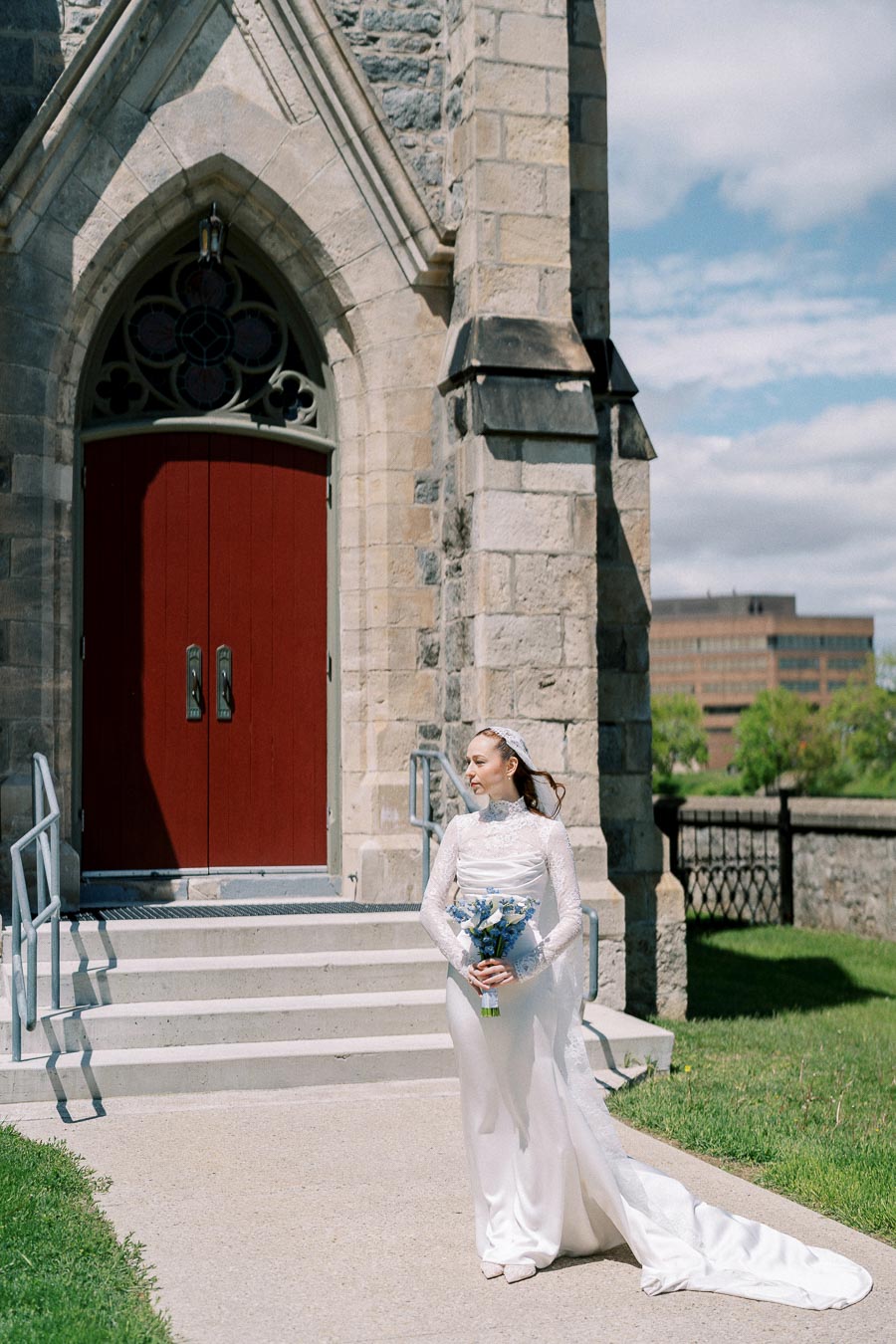 Bride in elegant white wedding dress and veil holding a bouquet of blue flowers, standing outside a stone church with red doors, under a clear sky.
