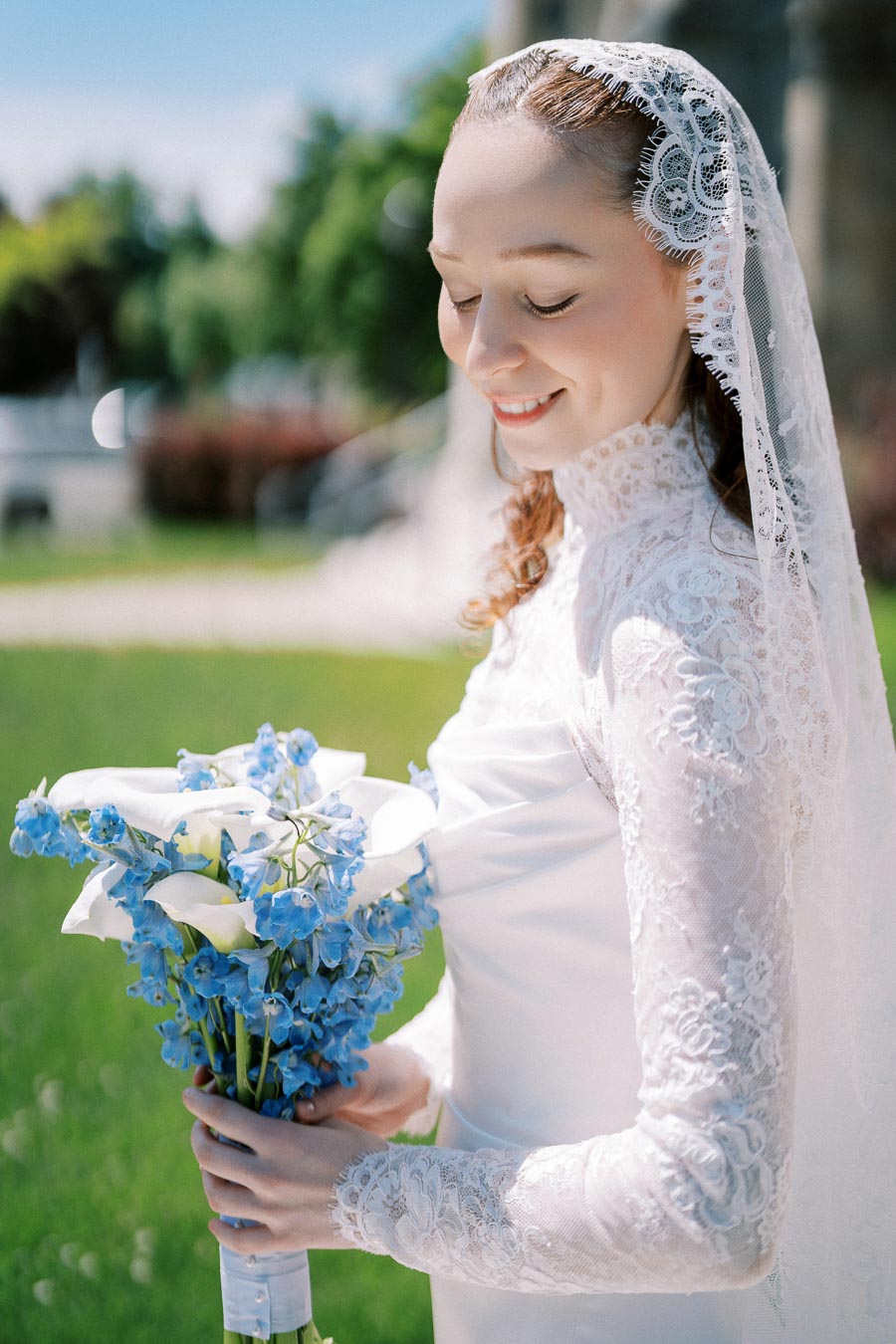 A smiling bride in a lace wedding dress and veil holding a bouquet of white calla lilies and blue flowers outdoors on a sunny day.