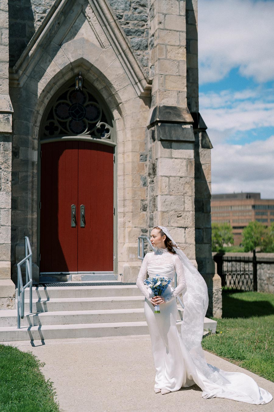 A bride in a white wedding dress holding a bouquet of blue flowers stands outside a historic stone church with red doors.