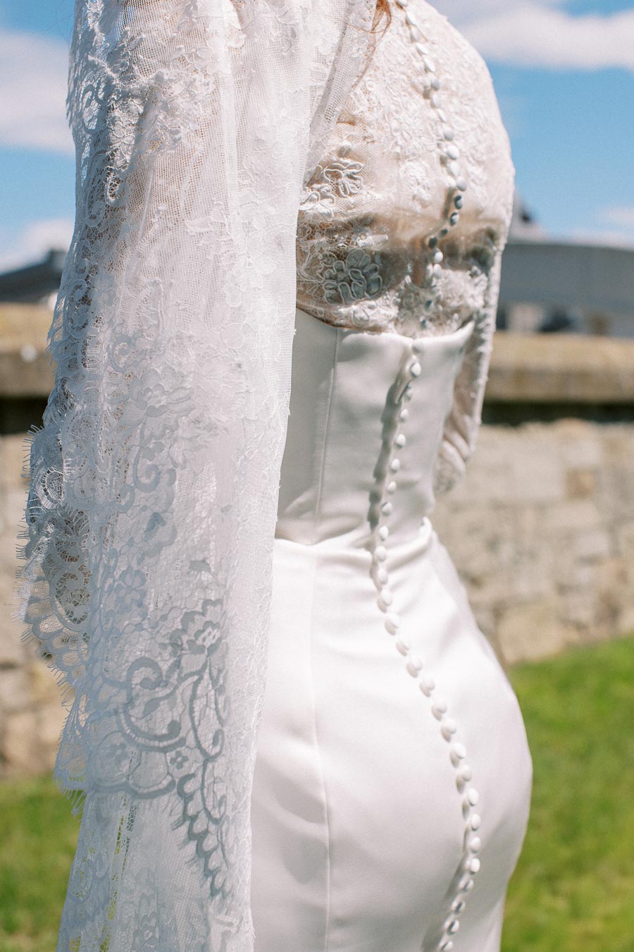 Close-up of a bride wearing an elegant lace wedding dress featuring intricate button detailing, set against a serene outdoor background on a sunny day.