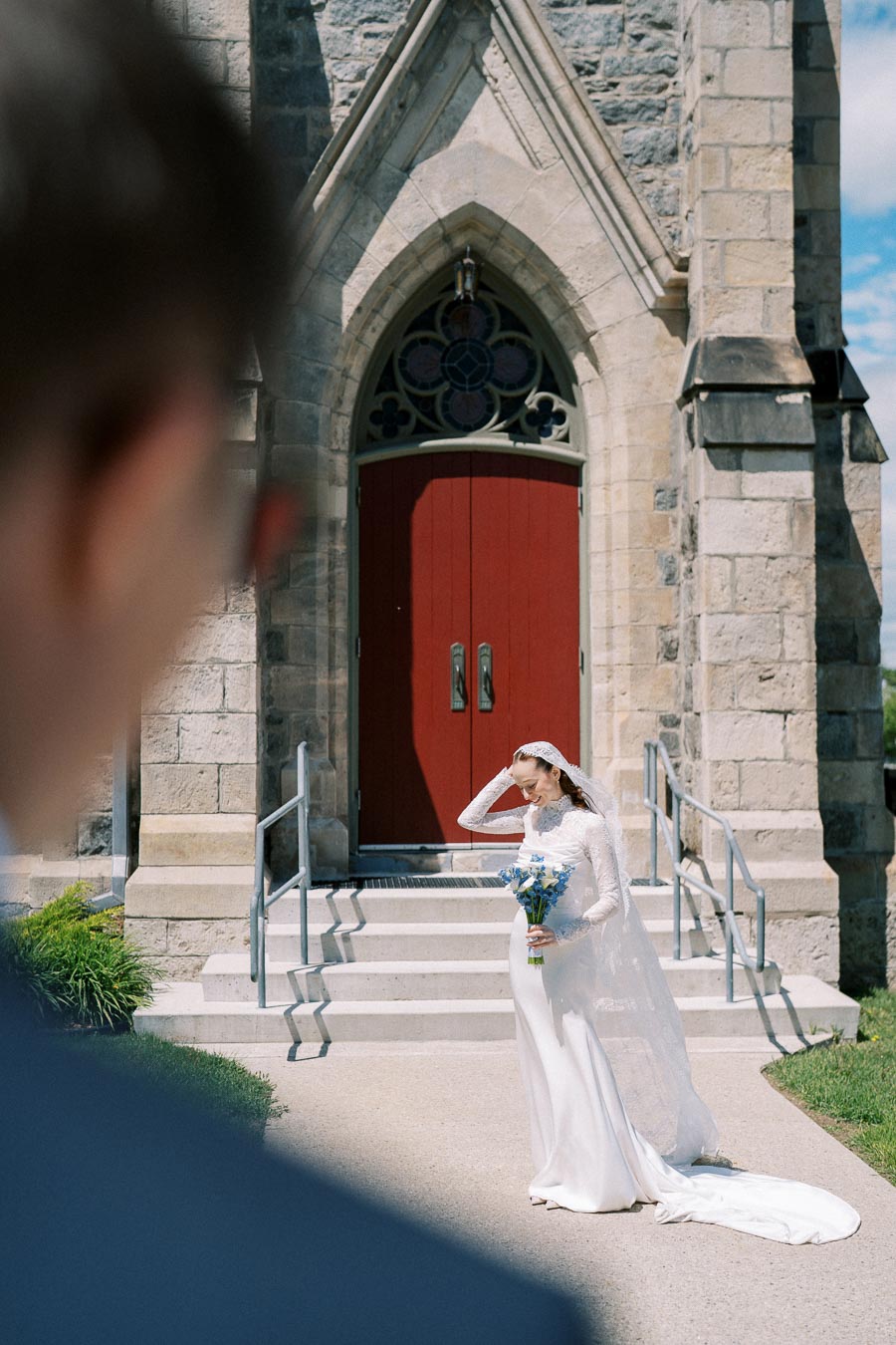 Bride in a long white dress holding blue flowers poses in front of a church with a red door on a sunny day, capturing a romantic pre-wedding moment.