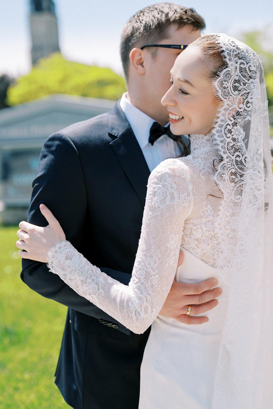A bride in a lace wedding dress and veil smiles as she embraces the groom in a dark suit and bow tie against a backdrop of greenery and blue sky.