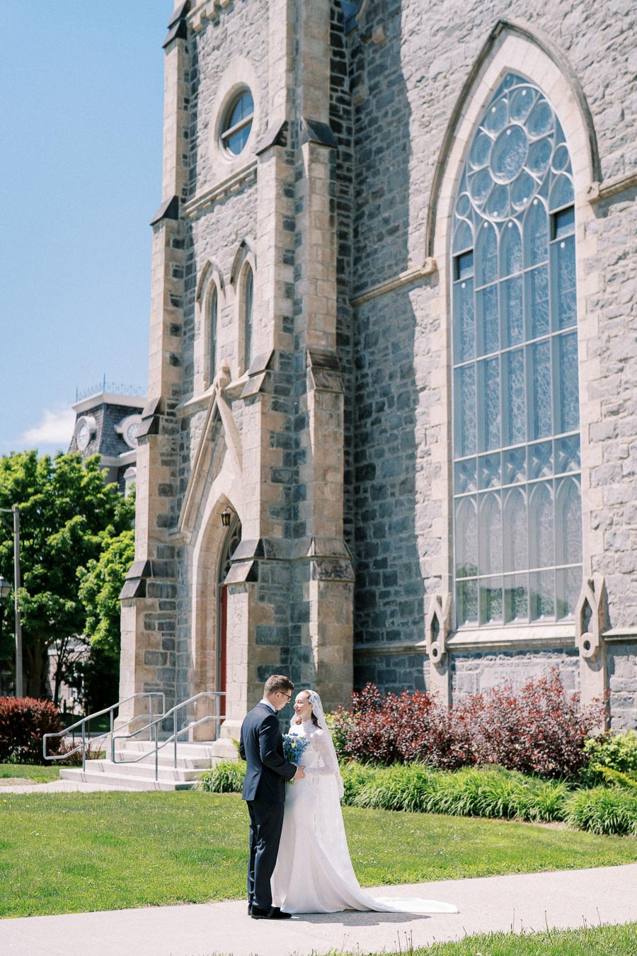 A bride and groom standing in front of a historic stone church, featuring tall stained glass windows, on a sunny day, with lush greenery around them.