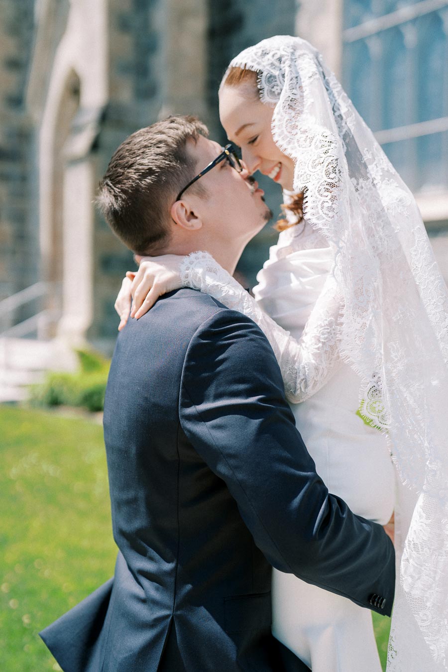 A bride in a lace veil embraces a groom in a navy suit outside a church.