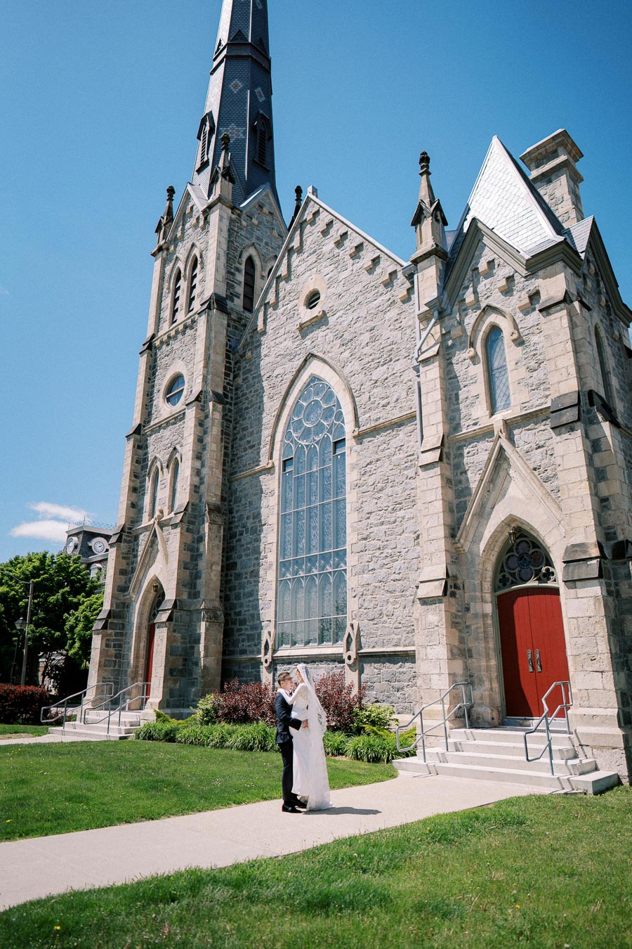 A happy couple embraces in front of a historic stone church with tall stained glass windows and red doors on a bright sunny day.