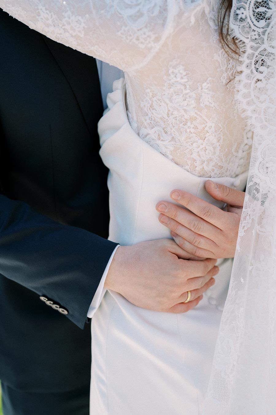 Close-up of a couple embracing on their wedding day, featuring the intricate lace of the bride's dress and the groom's dark suit. The groom's hand rests gently on the bride's waist, highlighting their wedding rings and the romantic moment.