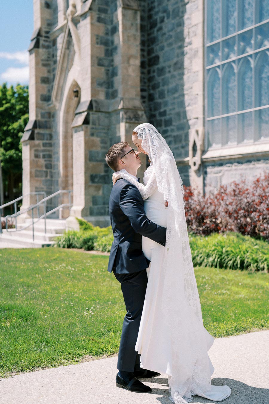 Bride and groom embracing joyfully in front of a historic church, wearing traditional wedding attire, with lush greenery and architectural details in the background.