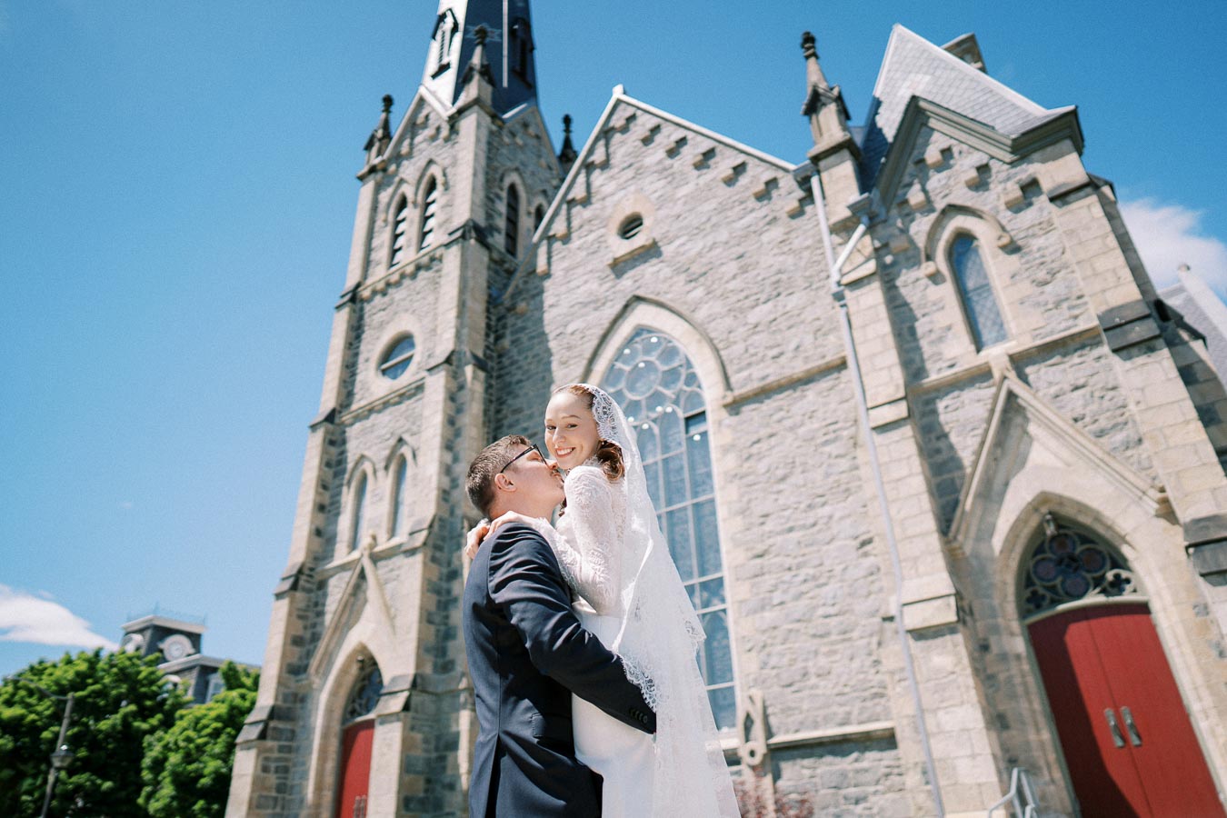 A groom lifting a smiling bride in a white lace wedding dress in front of a historic stone church with red doors under a clear blue sky.