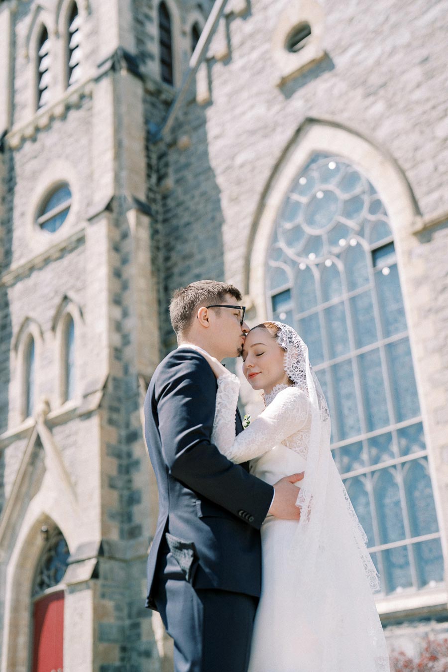 A bride and groom embrace lovingly in front of a historic stone church with large arched windows. The bride wears a white lace dress and veil, while the groom is dressed in a dark suit.