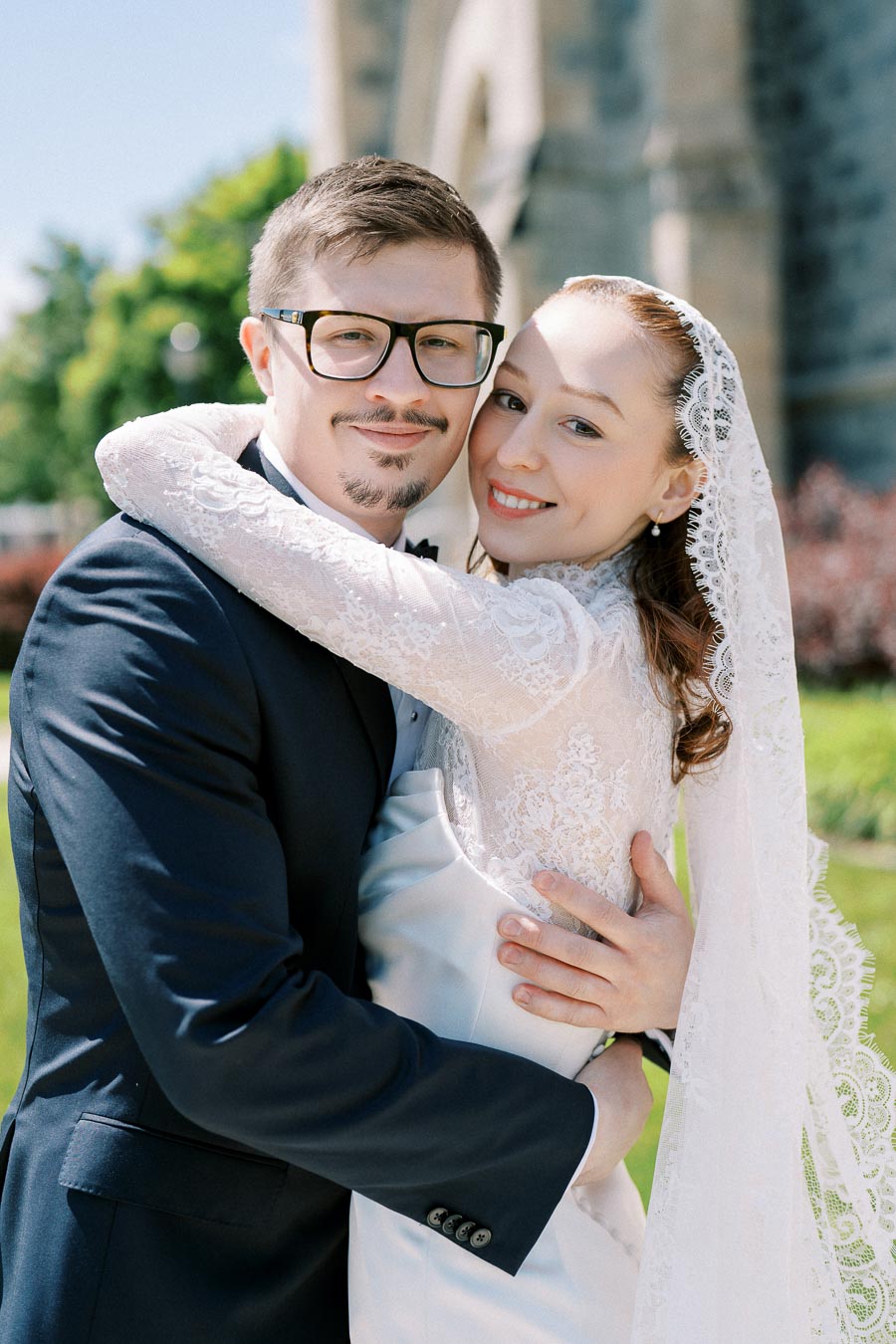 Happy bride and groom embracing outdoors, wearing formal wedding attire with lace details and glasses, in front of a scenic church backdrop.