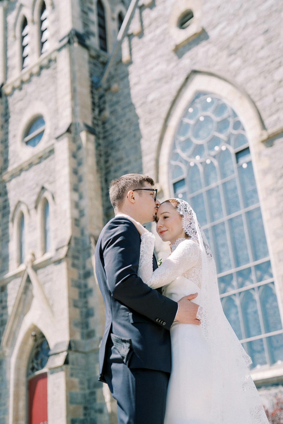 A couple embraces in front of a historic church with large stained glass windows, creating a romantic wedding scene under bright daylight.