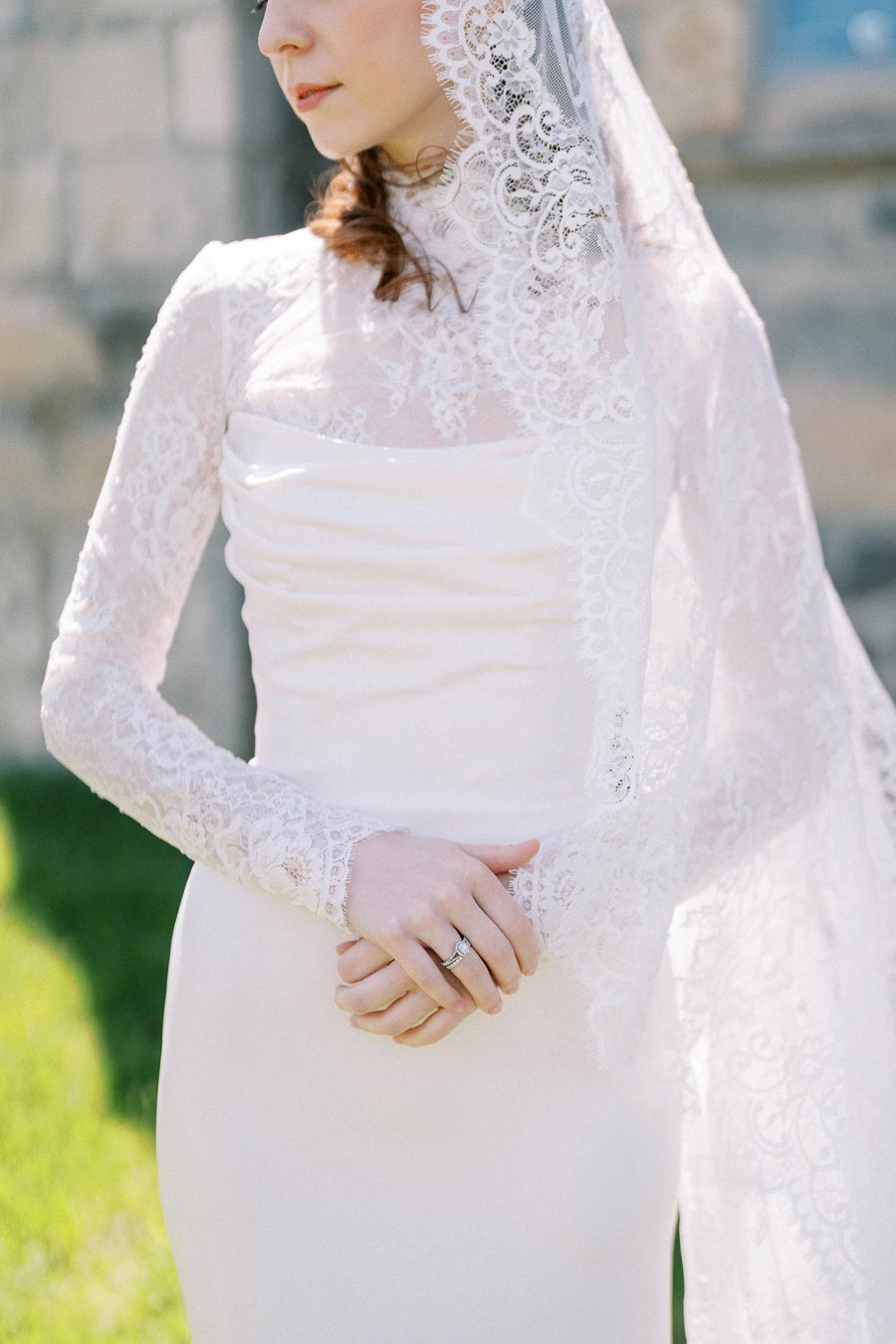 Elegant bride in lace wedding dress with intricate long sleeves and veil, displaying a delicate posture in a serene outdoor setting.