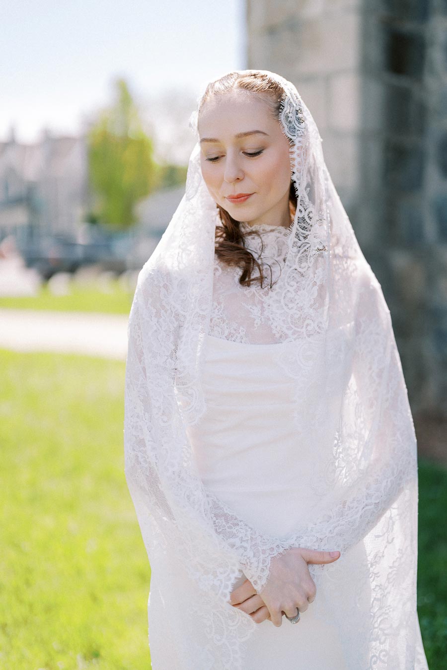 Elegant bride wearing a lace wedding veil outdoors on a sunny day, showcasing intricate lace detailing and serene expression.