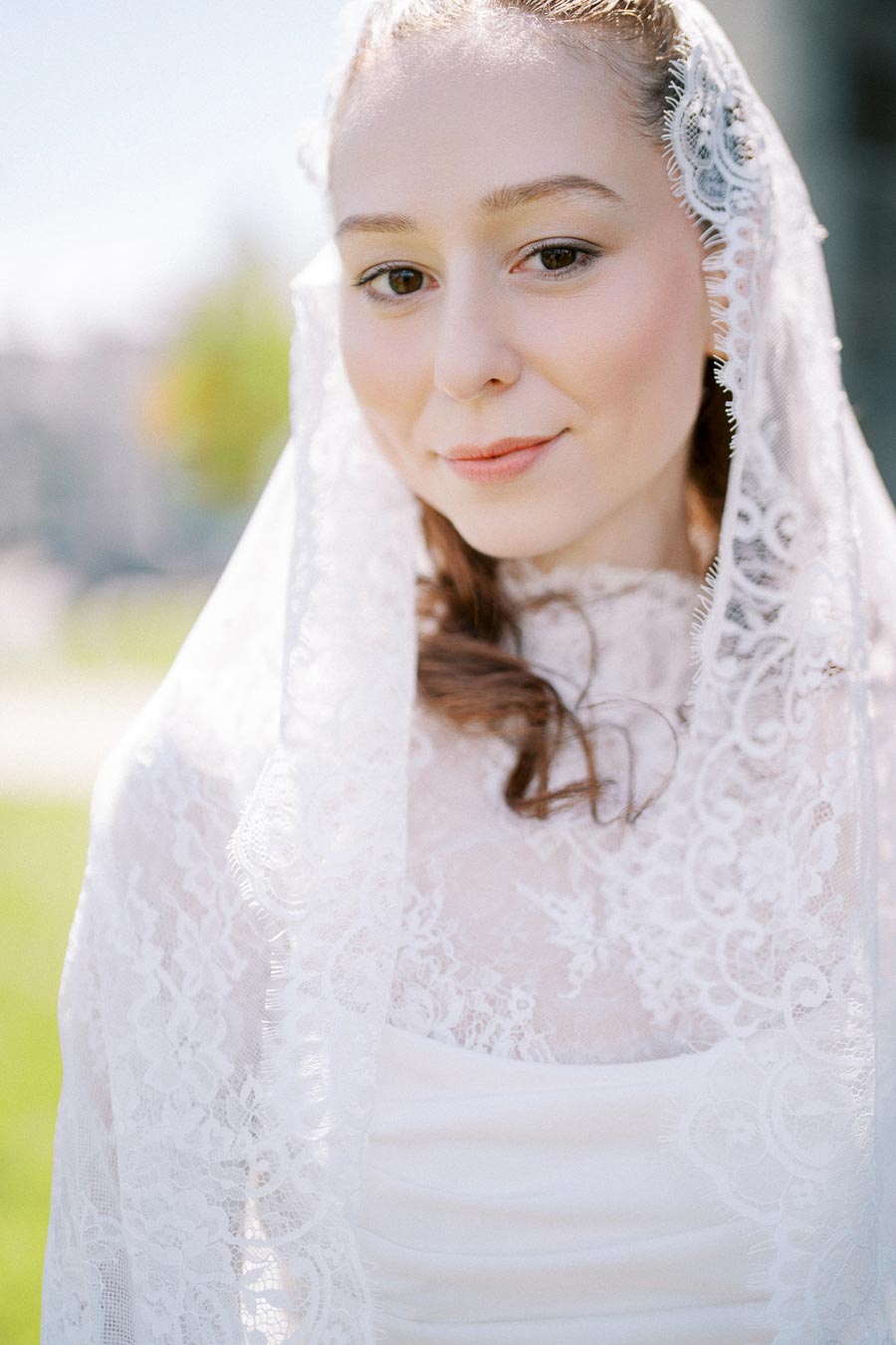 Young bride in a lace veil with a serene smile, posing outdoors under bright sunlight.