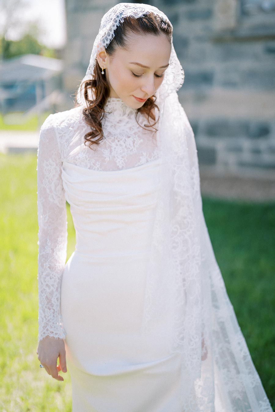 A bride in a delicate white lace wedding dress with a matching veil poses outdoors on a sunny day, showcasing elegant bridal fashion against a scenic background.