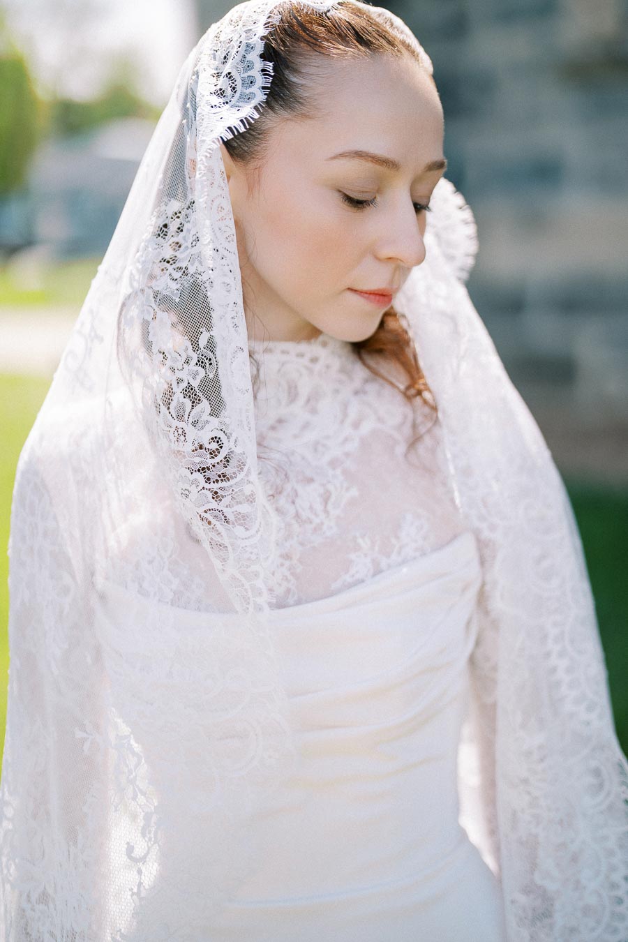A bride wearing an elegant lace wedding dress and veil, standing outdoors on a sunny day.