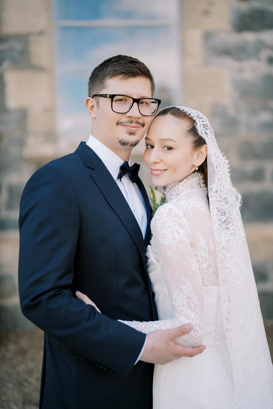 A bride and groom embracing on their wedding day, with the bride wearing a lace veil and the groom in a dark suit with glasses, standing in front of a stone building.