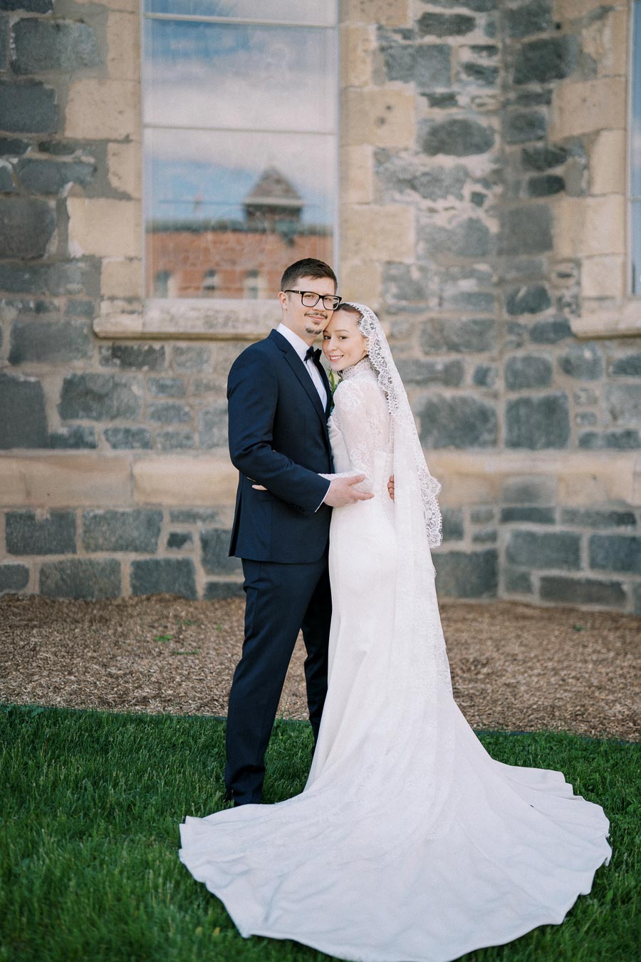 Elegant bride and groom posing outside a stone church wall, the bride in a lace wedding gown and veil, standing on green grass.