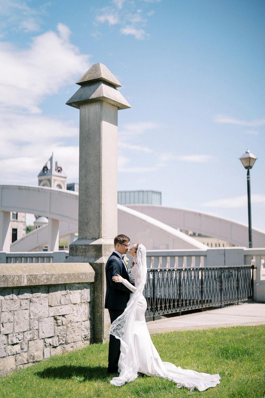 Romantic wedding couple embracing near a stone wall and bridge, with blue sky background on a sunny day.
