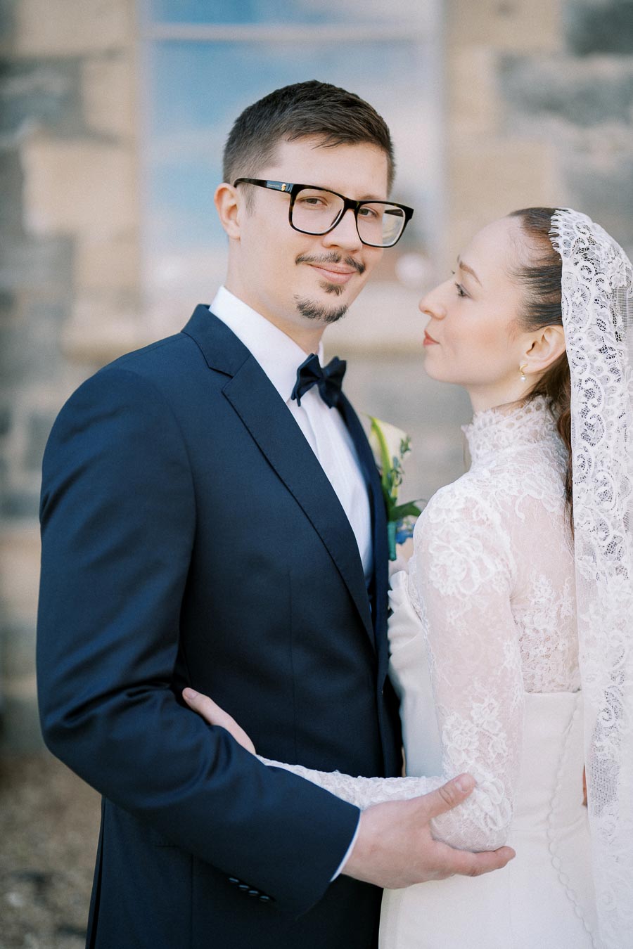 Bride and groom in elegant wedding attire pose in front of a stone building. The groom is wearing a dark suit with a bow tie, while the bride is in a lace gown with a veil.