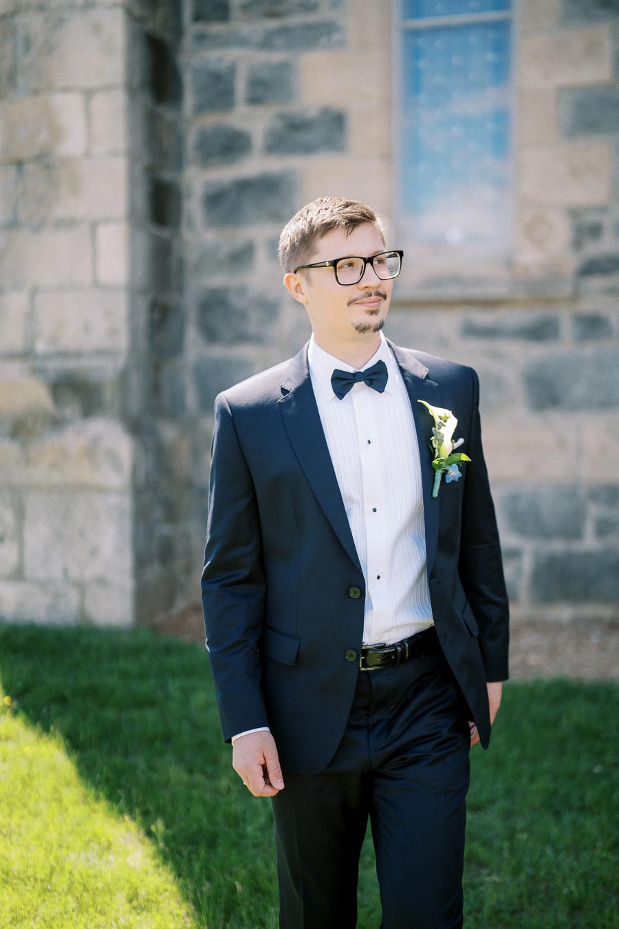 A man in a formal dark suit and bow tie, wearing glasses, standing confidently outside a stone building on a sunny day.