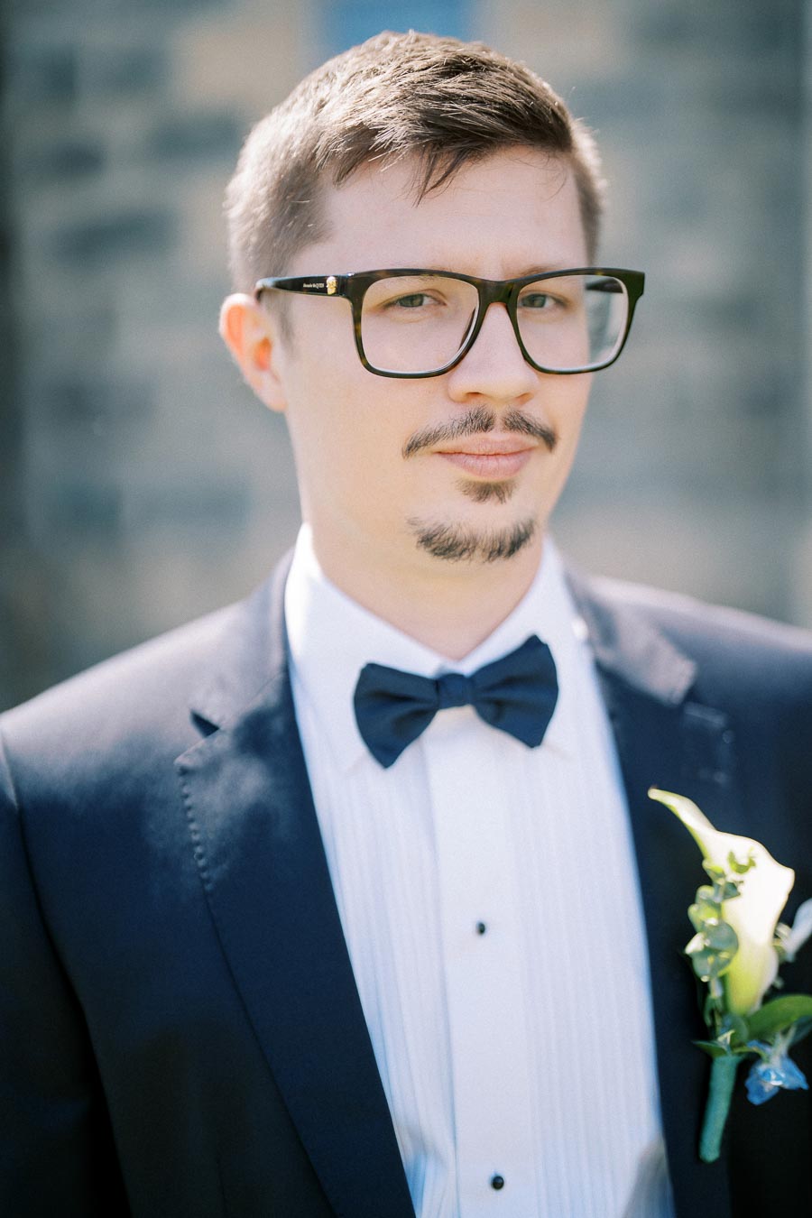 Young man in a formal dark suit and black bow tie with glasses and a boutonnière, standing outdoors with a blurred background.