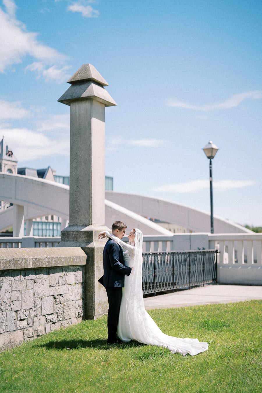 A bride and groom embrace on a sunny day beside a stone pillar and metal railing, with a modern bridge and clear blue sky in the background.