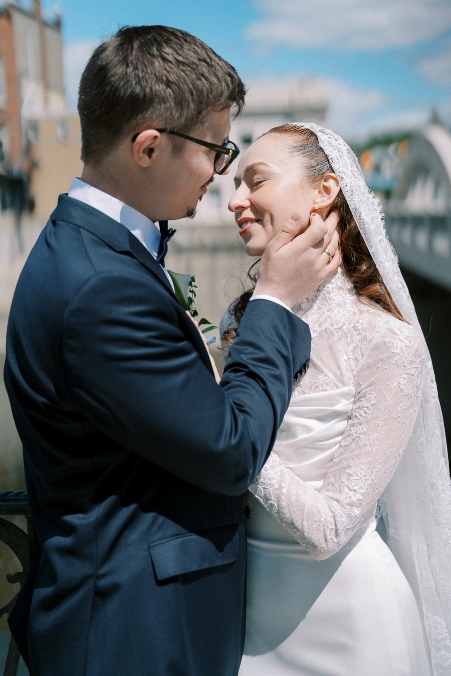 A bride and groom share an intimate embrace on a sunny day, with the groom gently holding the bride's face. The bride wears a lace veil and white dress, while the groom is in a dark suit. A bridge and blue sky provide a scenic backdrop.