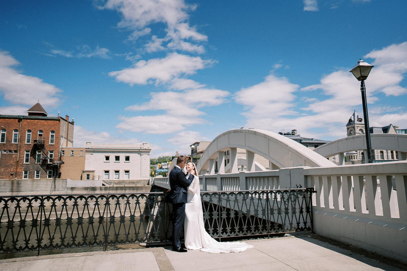 Couple embracing on a pedestrian bridge during a sunny day with architectural buildings and blue sky in the background.