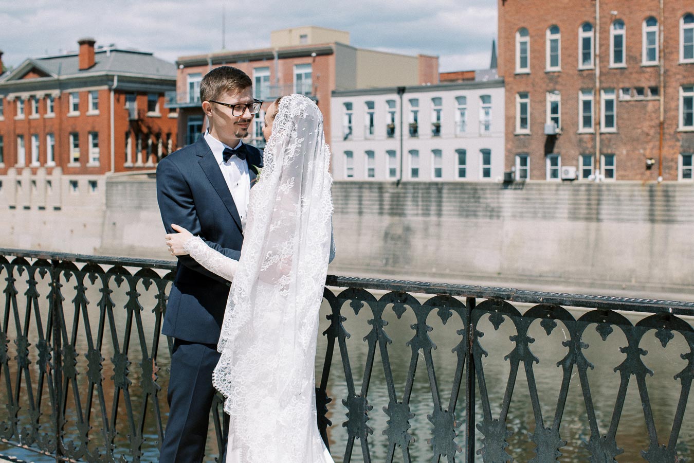 A bride and groom standing on a historic bridge in an urban setting, surrounded by old brick buildings, on a sunny day.